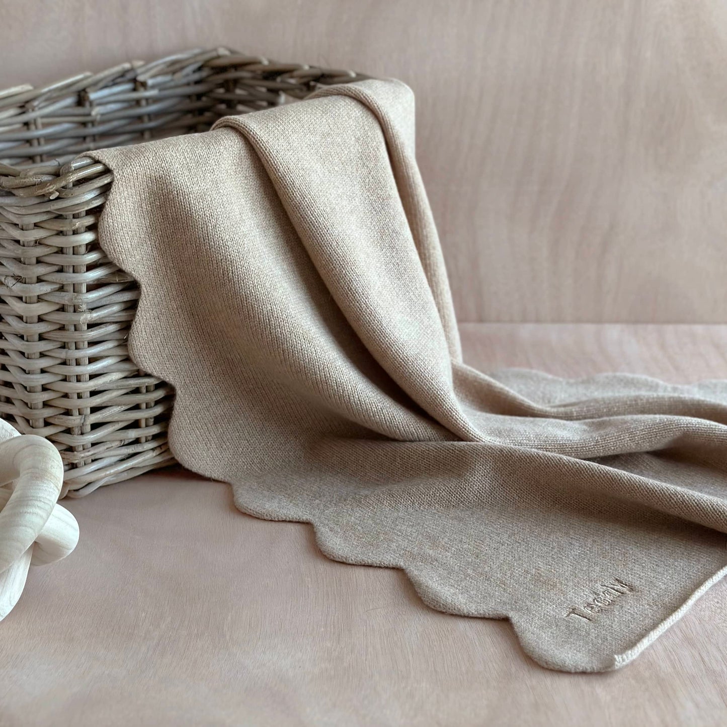Pure cashmere handmade baby blanket with scalloped edge in light beige colour draped over a wicker basket on a table.