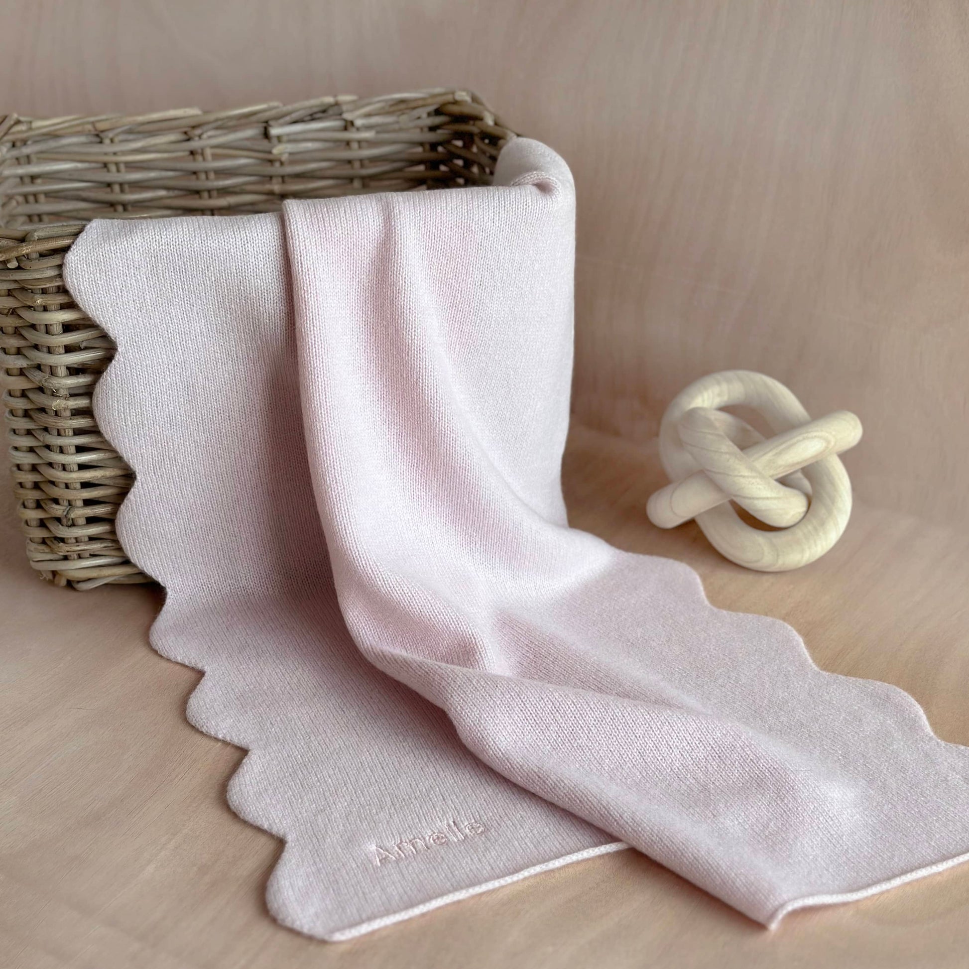 Pure cashmere new baby gift, scalloped edge baby blanket with personalisation draped over a wicker basket on a table.