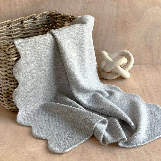 Pure cashmere handmade scalloped edge baby blanket in light grey colour with personalisation draped over a wicker basket on a table.